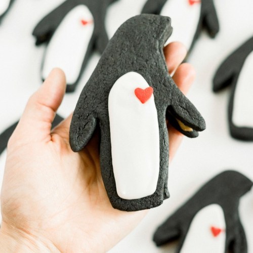 A hand holding a penguin-shaped cookie with white icing and a small red heart, surrounded by more cookies.