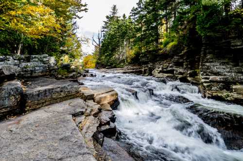 A flowing river surrounded by rocky banks and trees with autumn foliage under a partly cloudy sky.