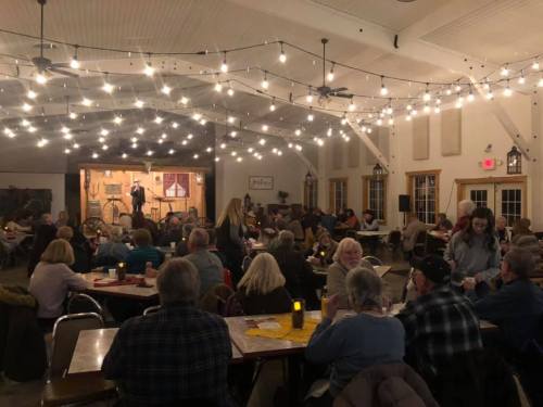 A lively indoor event with people seated at tables, string lights overhead, and a performer on stage.