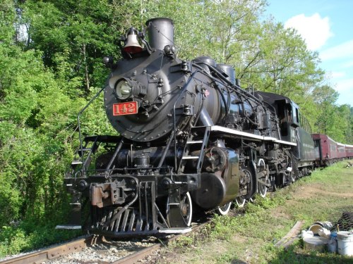 A vintage steam locomotive with a red number 142, parked on tracks surrounded by lush greenery.