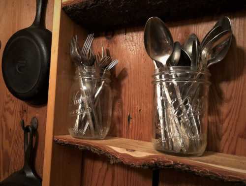 Two mason jars filled with silverware, including forks and spoons, on a wooden shelf against a rustic background.