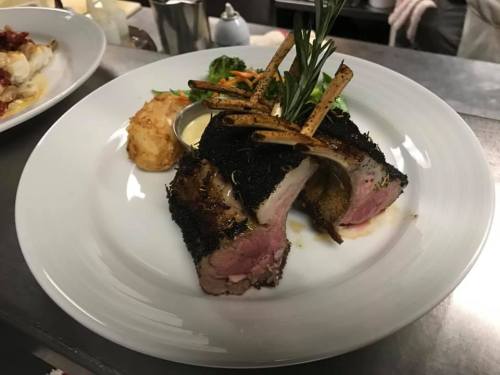 A beautifully plated rack of lamb with herbs, served with a side of vegetables and a creamy sauce.