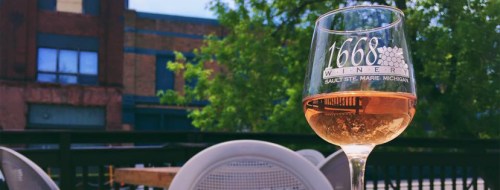 A glass of rosé wine sits on a table, with greenery and a building in the background.