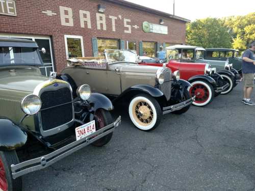 A row of vintage cars parked outside a brick building with "Bart's" signage.