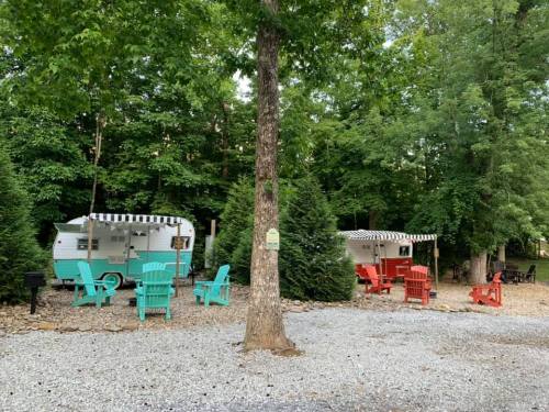 Two vintage campers in a wooded area, surrounded by colorful Adirondack chairs and gravel pathways.