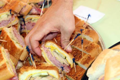 A hand reaching for a sandwich with toothpicks, surrounded by several other sandwiches on a platter.
