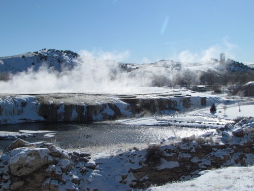 A snowy landscape with steam rising from a river, surrounded by hills and a clear blue sky.