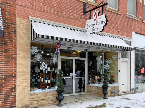 A cozy tea shop with a striped awning, festive decorations, and snowflakes on the windows, located in a brick building.
