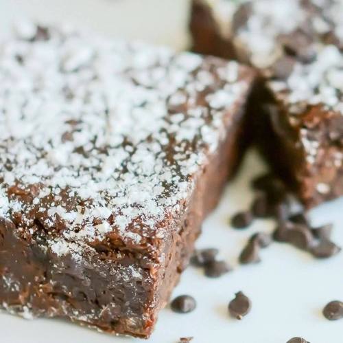 Close-up of rich chocolate fudge topped with powdered sugar and scattered chocolate chips on a white plate.