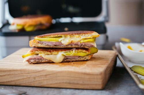 A grilled sandwich with layers of meat, cheese, and pickles, served on a wooden cutting board.