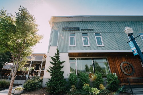 Modern building with green metal siding, large windows, and surrounding greenery, under a bright blue sky.