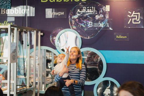 A woman smiles while holding a baby, surrounded by colorful bubbles in a playful indoor setting.