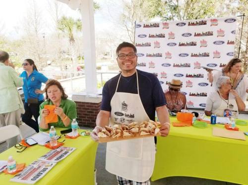 A smiling man in an apron holds a tray of food, while others enjoy snacks at a lively outdoor event.