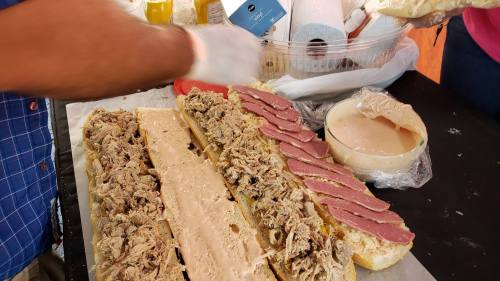 A person prepares a large sandwich with layers of pulled meat and sliced meat on a long bread roll.