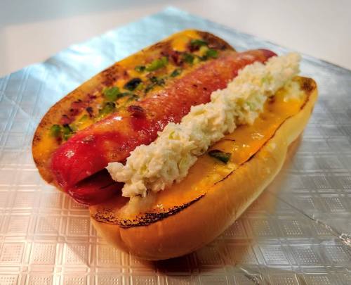 A grilled hot dog in a toasted bun, topped with creamy coleslaw and jalapeños on a silver foil background.