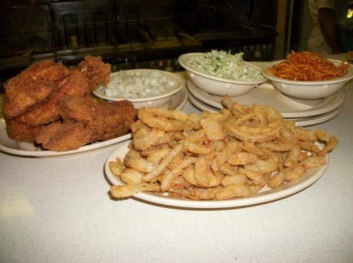 A variety of fried foods on plates, including crispy fried chicken, onion rings, and side dishes like coleslaw and rice.