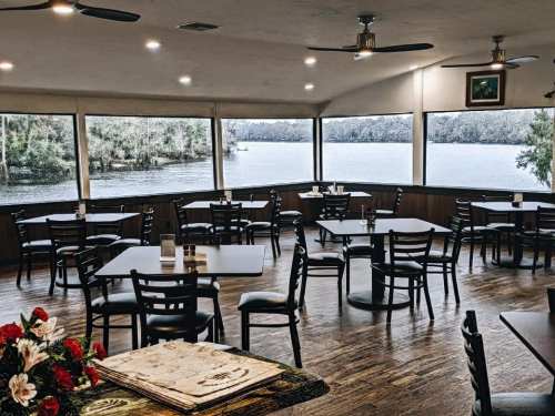 A cozy restaurant interior with empty tables, large windows overlooking a scenic water view, and floral decor.
