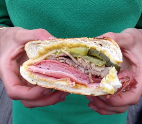 A person holding a sandwich filled with layers of meats, pickles, and mustard, with a green background.
