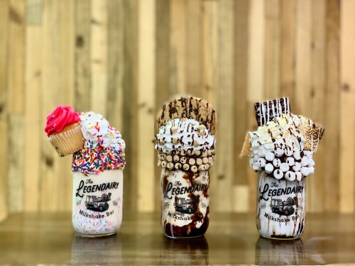 Three extravagant milkshakes in jars, topped with colorful sprinkles, whipped cream, and various treats, against a wooden backdrop.