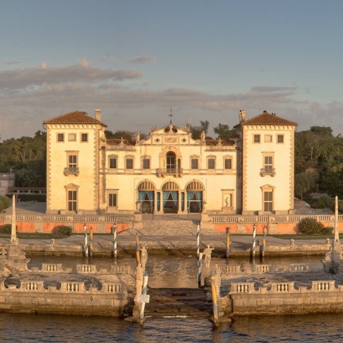 Historic mansion with ornate architecture, surrounded by water and lush greenery, under a clear blue sky.