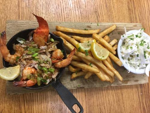 A cast iron skillet with shrimp and lobster, served with fries and coleslaw, garnished with lemon slices.