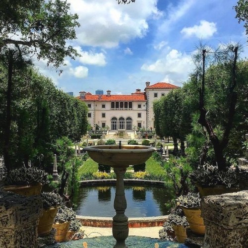 A grand mansion surrounded by lush gardens and a fountain, under a bright blue sky with fluffy clouds.