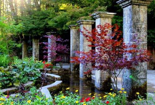 A serene garden scene featuring stone columns, vibrant flowers, and colorful trees beside a tranquil pond.
