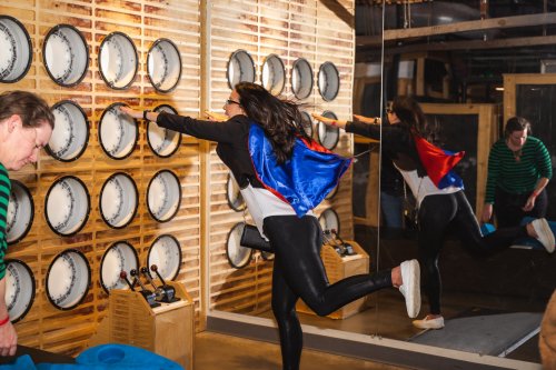 A person in a cape playfully interacts with a wall of circular instruments, while another person observes nearby.