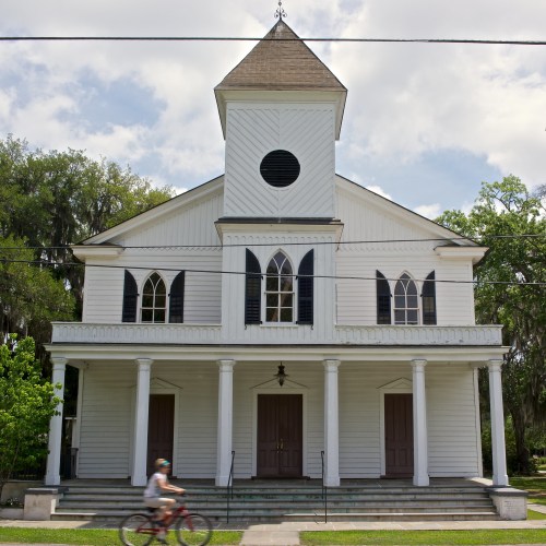 A white church with a tall steeple and columns, featuring large windows and a cyclist passing by in front.