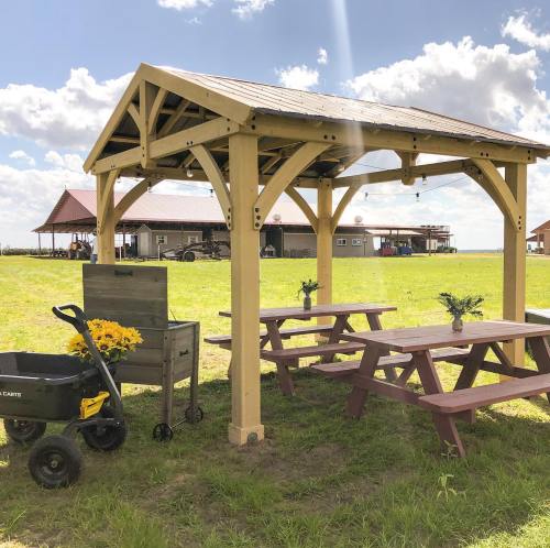 A wooden gazebo with picnic tables and a cart of flowers in a grassy field under a blue sky with clouds.