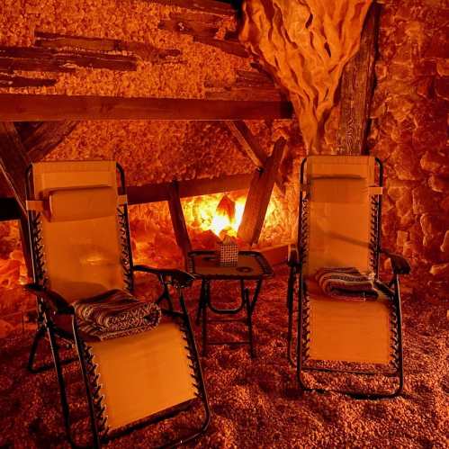 Two lounge chairs with blankets sit beside a small table in a warm, salt cave setting, illuminated by soft orange light.