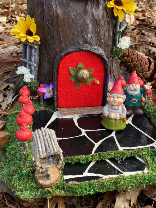 A whimsical fairy house with a red door, surrounded by flowers and gnome figurines on a mossy base.