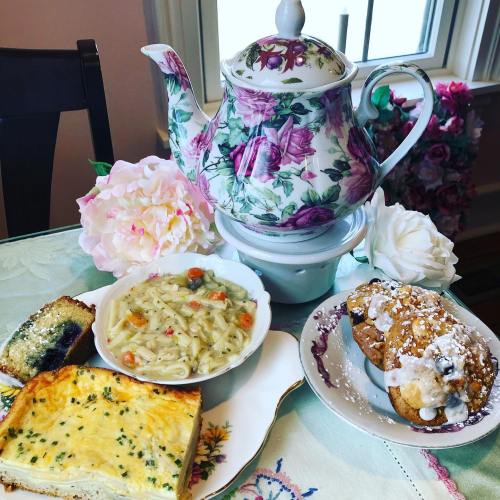 A floral teapot with a plate of soup, quiche, and pastries, surrounded by pink flowers on a table.