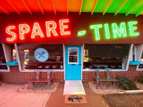 Bright neon sign reading "SPARE-TIME" above a turquoise door, with benches outside a cozy storefront.