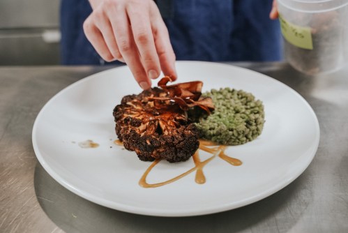 A hand garnishes a plate with a charred cauliflower steak, green puree, and a drizzle of sauce.