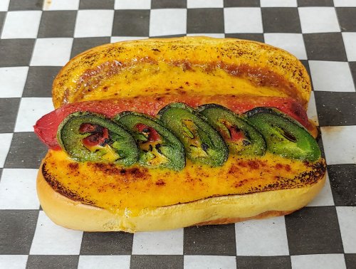 A grilled hot dog topped with melted cheese and sliced jalapeños on toasted bread, set on a checkered background.