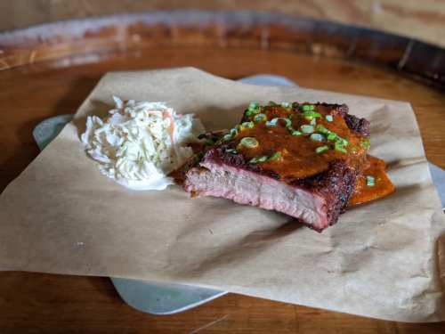 A slice of smoked brisket topped with sauce and green onions, served with a side of coleslaw on brown paper.