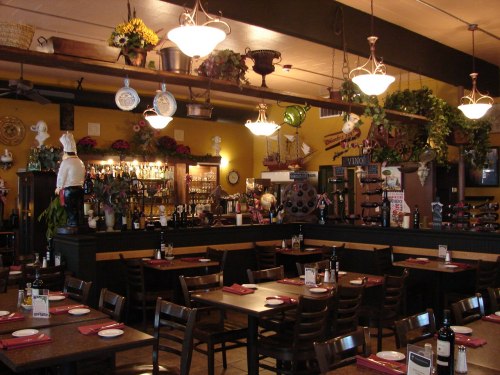 Cozy restaurant interior with wooden tables, wine bottles, and decorative plants, featuring a bar and warm lighting.