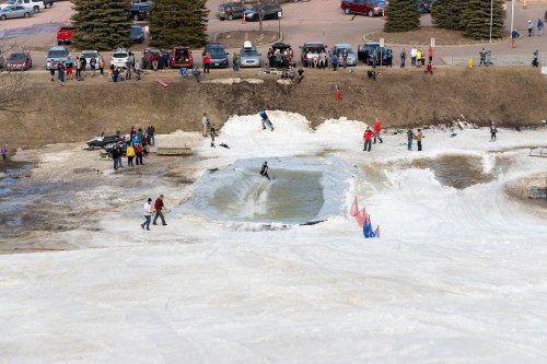 A snowy hill with people skiing and sliding into a water pool, surrounded by spectators and parked cars.