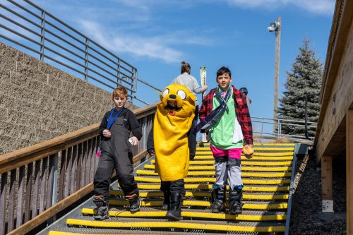 Three kids walk down outdoor stairs; one wears a bright yellow cartoon character costume, while others are in winter gear.