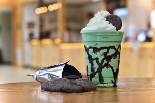 A mint green milkshake topped with whipped cream and chocolate drizzle, next to a bag of cookies and some cookies on a table.