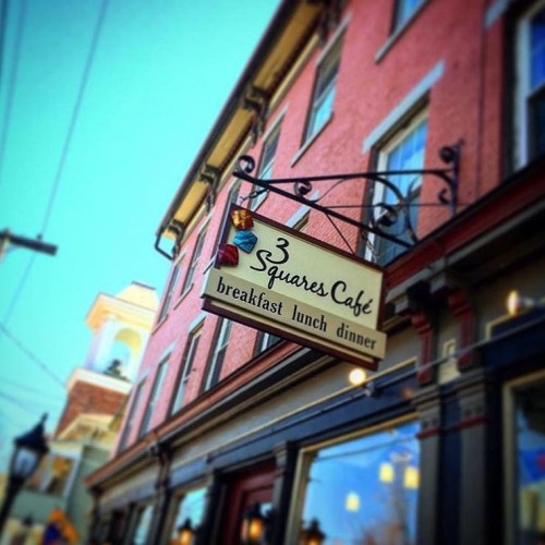 Sign for 3 Squares Cafe hanging outside a pink building, advertising breakfast, lunch, and dinner.