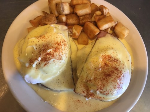 A plate of eggs Benedict topped with hollandaise sauce and paprika, served with diced, golden-brown potatoes.