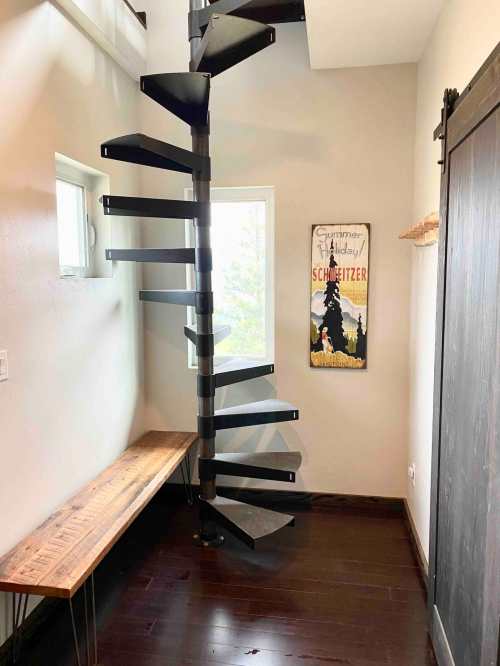 A modern spiral staircase leads to a bright window, with a wooden bench and a vintage poster on the wall.