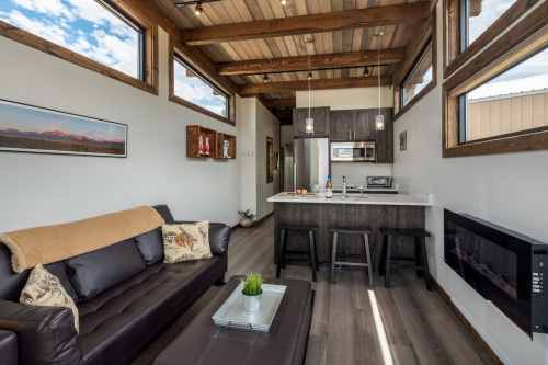 Cozy modern living space with a leather sofa, wooden beams, and a kitchen area featuring bar stools and natural light.