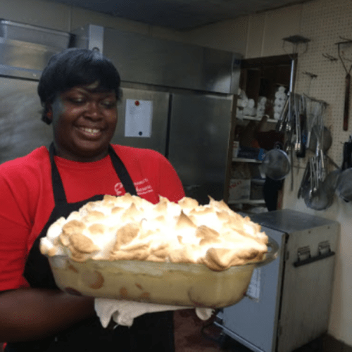 A smiling person in a red shirt holds a large dish of meringue-topped dessert in a kitchen setting.