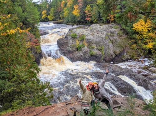 Two people pose joyfully by a rushing river surrounded by colorful autumn foliage.