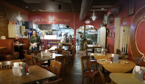 Cozy restaurant interior with wooden tables, chairs, and a counter in the background; a server is attending to customers.