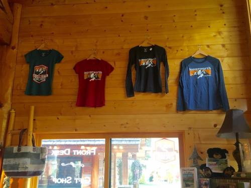 Four t-shirts hanging on a wooden wall, featuring various designs and colors, in a cozy shop setting.