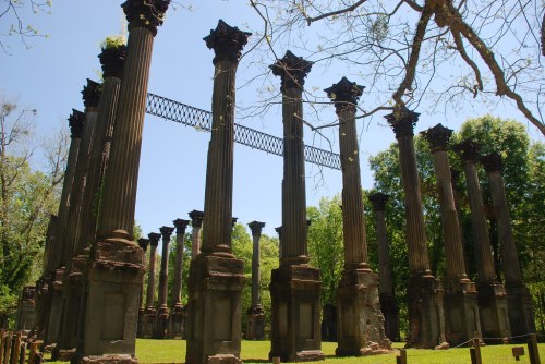 Ruins of tall columns in a green landscape, with trees and a clear blue sky in the background.